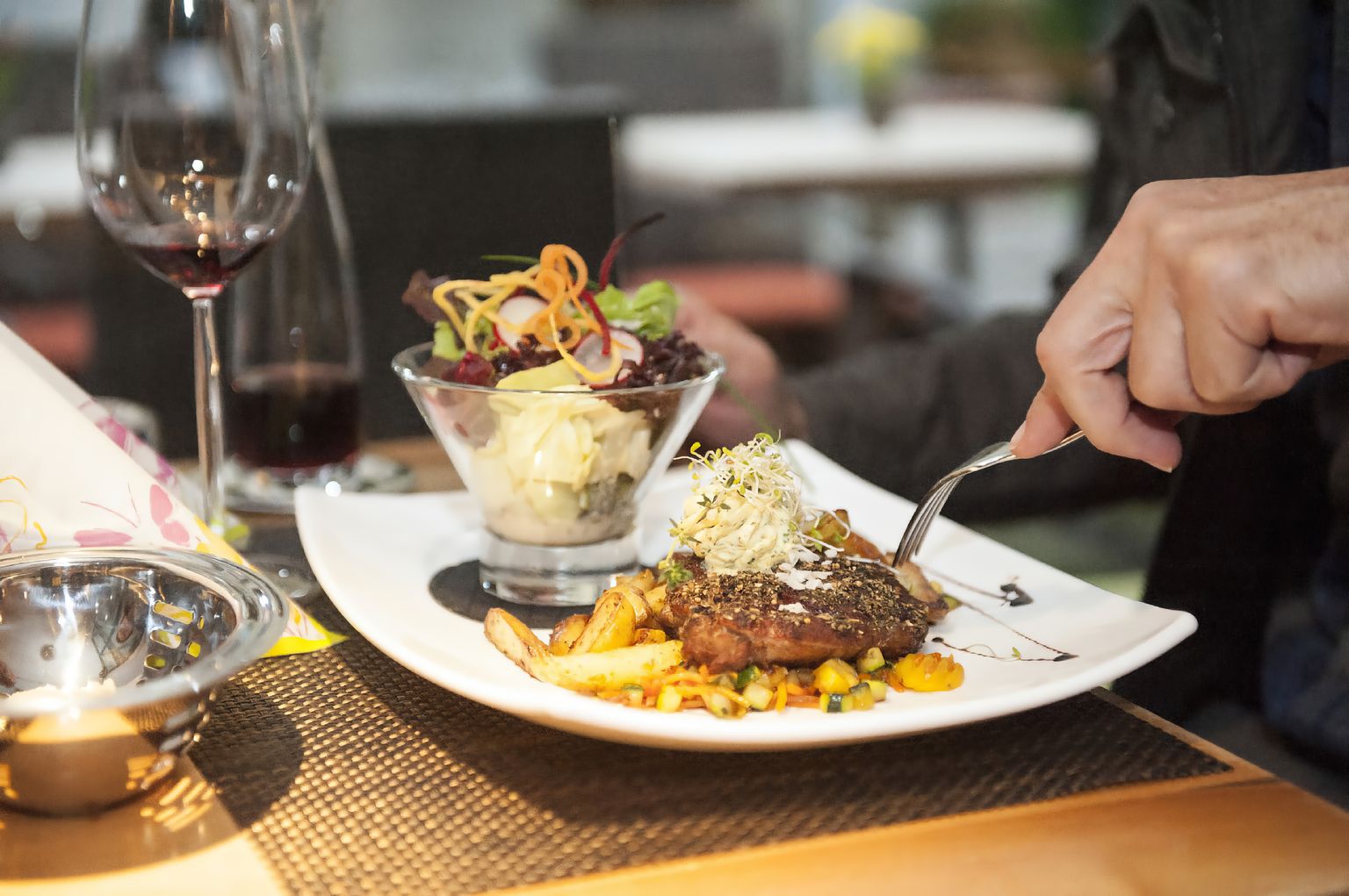 Ein rechteckiger Teller, belegt mit einem garnierten Entrecôte, Grillgemüse und Pommes frites neben einem bunten Salatglas, auf einem Tisch zusammen mit einem Glas Rotwein und der Weinkaraffe.
